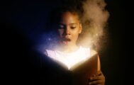 A young Black girl gasps in surprise as she reads. Her book is glowing, as if by magic, and lights up her face.